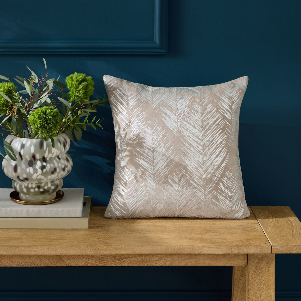 Folius Velvet Cotton Cushion in silver, a 43 x 43cm square with a foil-printed herringbone leaf pattern, shown on a wooden bench against a dark blue wall.