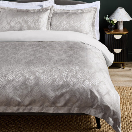 Folius Cotton Leaf Duvet Cover Set in Silver with matching pillowcases, shown on a bed with white trim, featuring a jacquard leaf pattern in a bedroom setting with a dark bedside table and lamp.