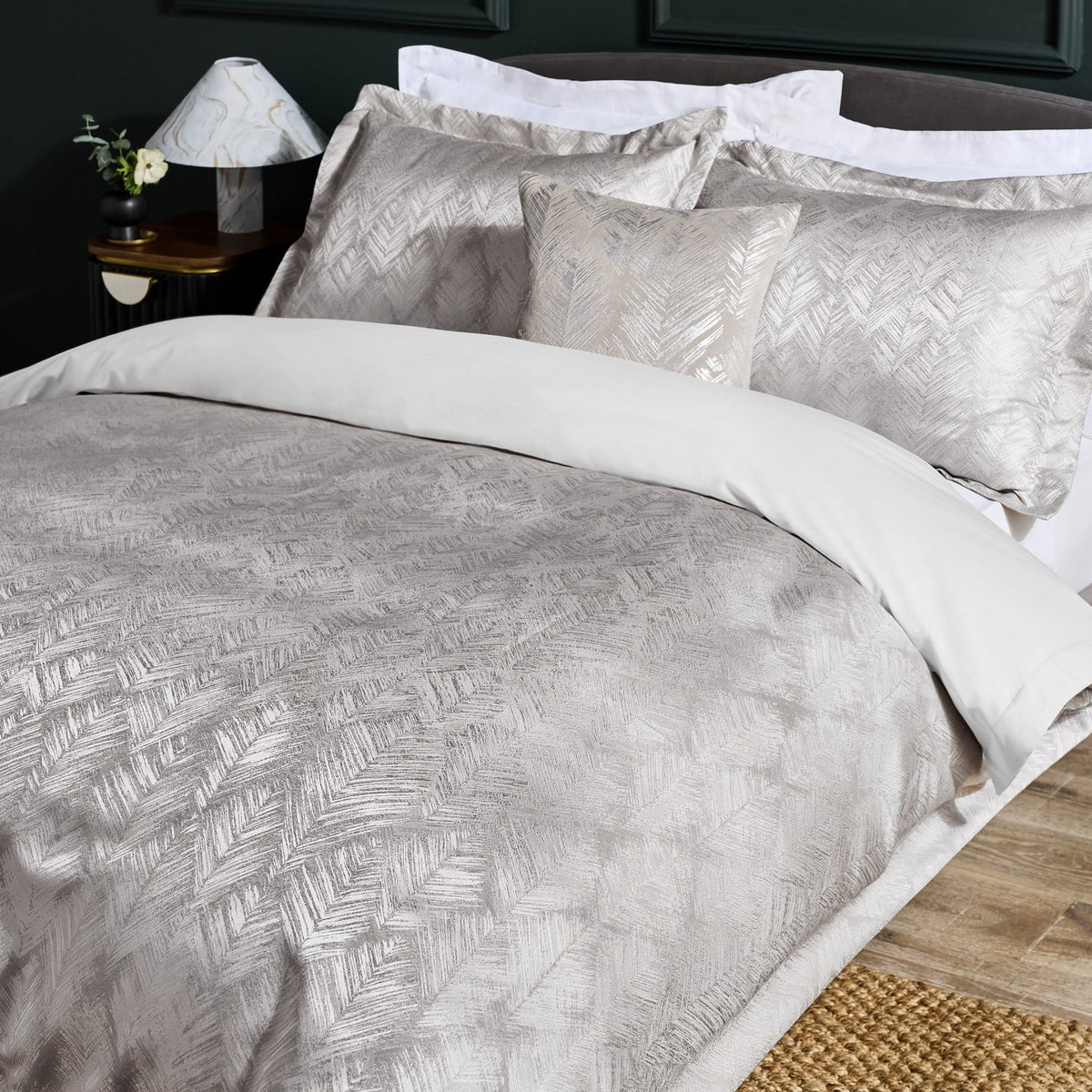 Folius Cotton Leaf Duvet Cover Set in Silver dressed on a bed with matching pillowcases and a cushion, styled in a bedroom with a dark headboard and bedside lamp.