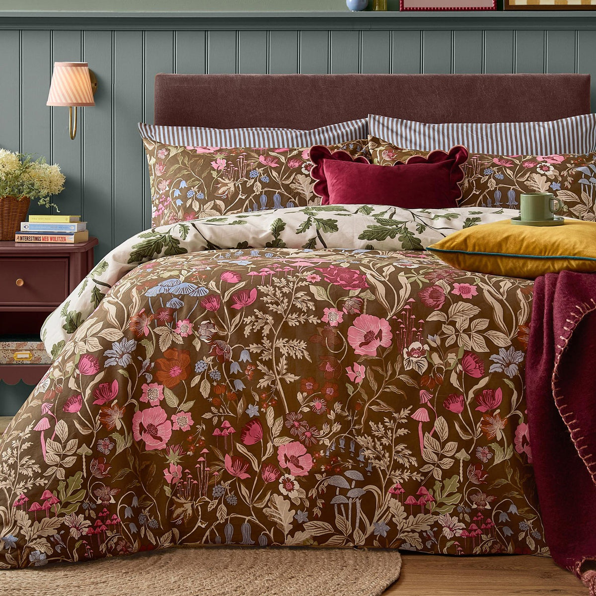 Chocolate Forest Bloom reversible duvet set with floral, wildflower and mushroom print, cream reverse, striped pillowcases and burgundy velvet cushions on a bed.