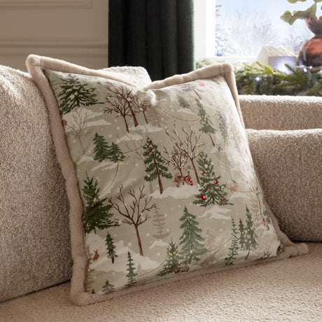 Forest Festivities Velvet Faux Fur Cushion in natural, square with printed winter woodland scene and faux fur trim, shown on a sofa in a living room.
