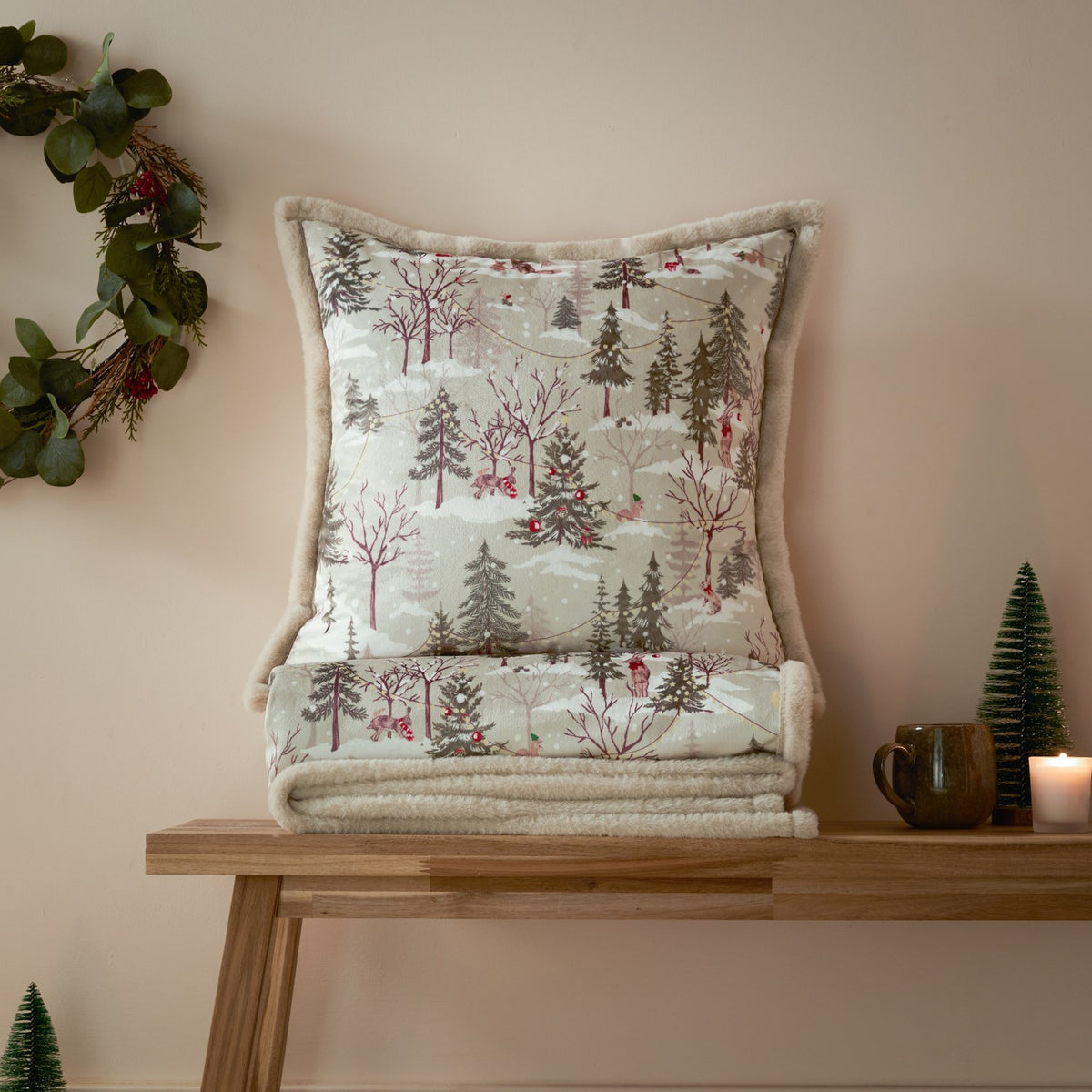 Forest Festivities Velvet Faux Fur Cushion in natural, shown with matching folded throw on a wooden bench, featuring a printed winter woodland scene with pines, animals and cream faux fur trim.