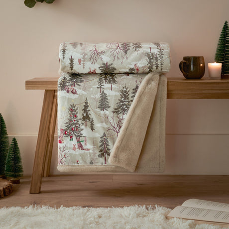 Forest Festivities Velvet Faux Fur Throw in natural, shown rolled over a wooden bench, displaying a woodland print face and cream faux fur reverse in a festive living room setting.