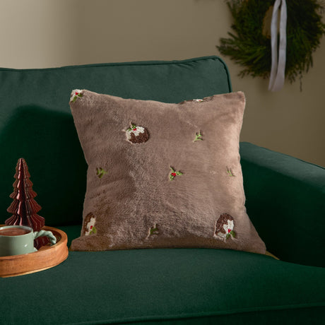 Square faux fur cushion in mocha brown with embroidered Christmas pudding and holly motifs, shown on a dark green sofa in a festive living room setting.