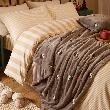 Festive Puddings Velvet Faux Fur Throw in mocha brown draped over a bed with striped bedding, showing embroidered Christmas pudding motifs with holly detail.