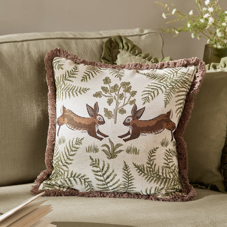 Fable Rabbits Woodland Cotton Cushion in willow green, square with fringe trim, showing two leaping rabbits and fern motifs, styled on a sofa.