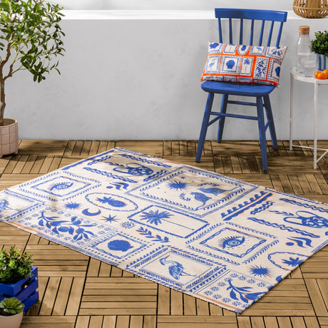 Frieze Abstract Polyester Rug in blue, cream flatweave with folk-art motifs including herons, palms and eye designs, shown on decking beside a blue chair.