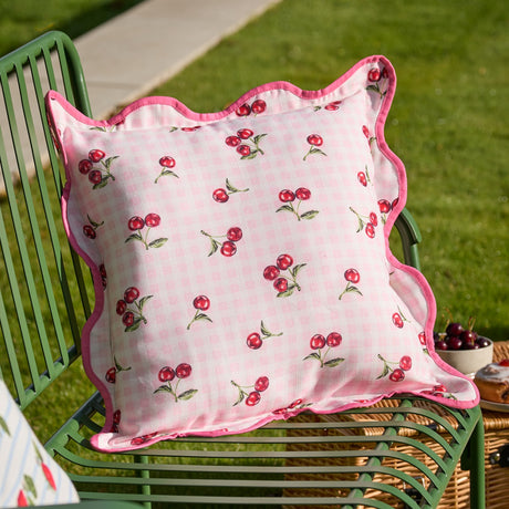 Square Fruits Gingham Polyester Cushion in pink, printed with red cherries on gingham check, scalloped edge with pink piped trim, shown on a green garden chair outdoors.