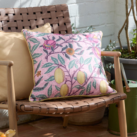 Fruits Piped Polyester Cushion in Blush Pink/Lemon with lemon and pomegranate print, yellow piped edge, shown on a woven outdoor bench.