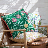 Fruits Piped Polyester Cushion in coral pink and lemon, with pomegranate and citrus print cushions on a wooden chair against a white brick wall.