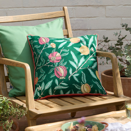 Fruits Piped Polyester Cushion in Forest Green, 45x45cm, with pink pomegranates and golden fruit print, coral piped trim, shown on a wooden garden bench beside a plain green cushion.