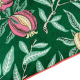 Close-up of the Fruits Piped Polyester Cushion in Forest Green, showing pink pomegranates, golden fruits and sage leaves on a green ground with a coral-orange piped edge trim.