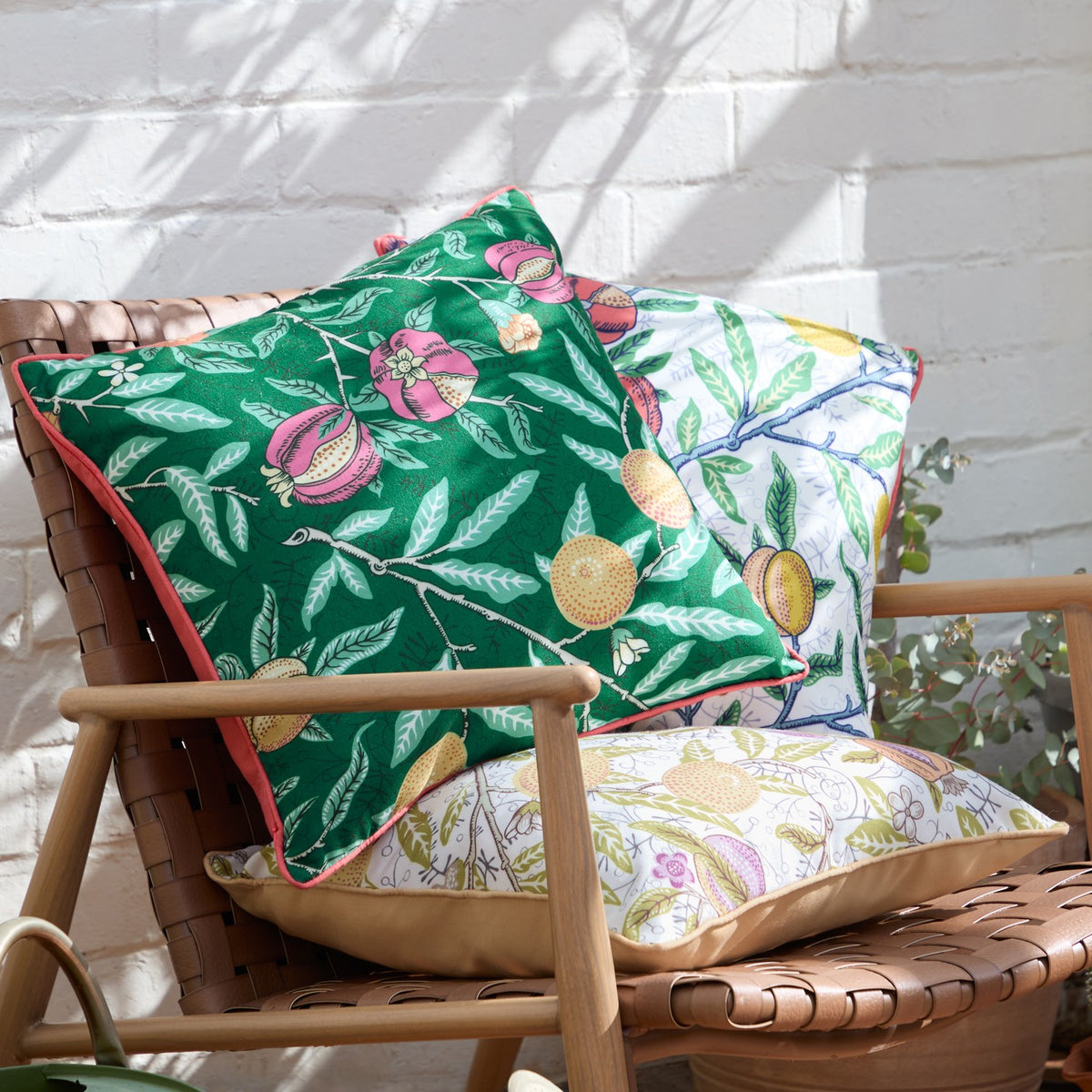 Fruits Piped Polyester Cushion in Forest Green with pomegranate and floral print, coral piped edge, styled on a woven chair beside a matching white botanical-print cushion.
