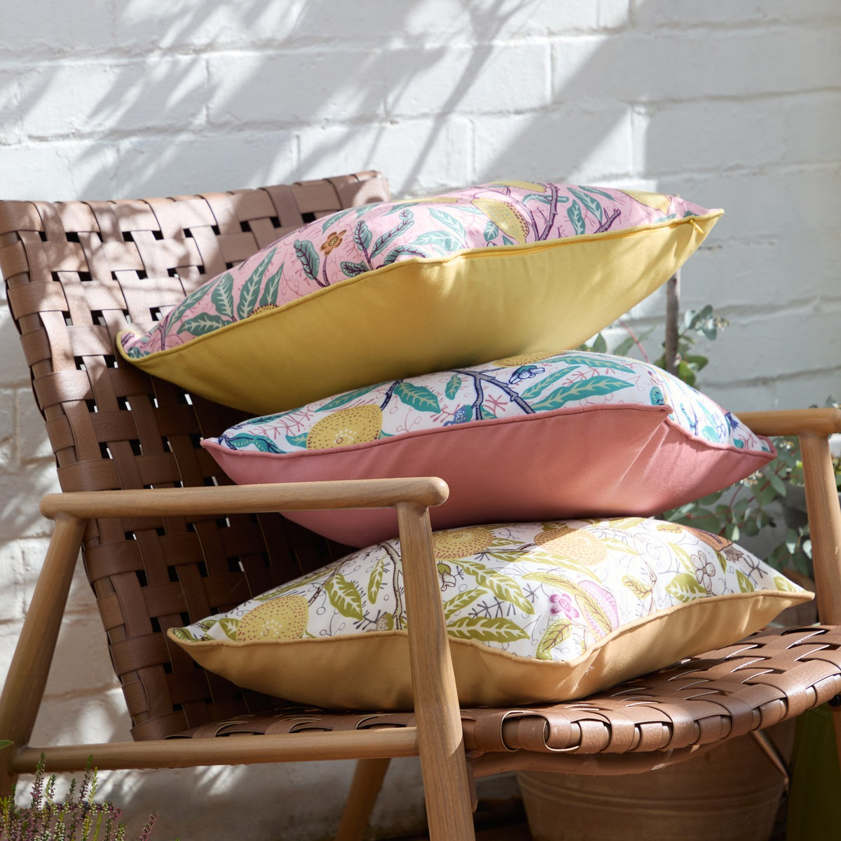 Three Fruits Piped Polyester Cushions stacked on a wooden garden chair, each with cream fronts printed in pink, green and yellow floral fruit patterns, plain yellow or pink piped reverse sides.