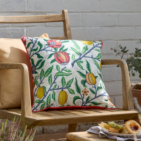 Fruits Piped Polyester Cushion in Yellow/Tomato Red, a square outdoor cushion with pomegranates, lemons and foliage on a pale ground with red piping, shown on a wooden garden bench.