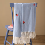 Fruits Knitted Polyester Throw in Berry Sweet Blue draped over a blue chair, featuring embroidered red strawberry motifs and cream fringed edges.