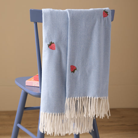 Fruits Knitted Polyester Throw in Berry Sweet Blue draped over a blue chair, featuring embroidered red strawberry motifs and cream fringed edges.