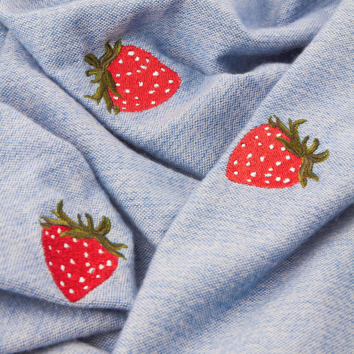 Fruits Knitted Polyester Throw in Berry Sweet Blue, close-up of chambray-blue fabric with embroidered red strawberry motifs and green calyx detailing.