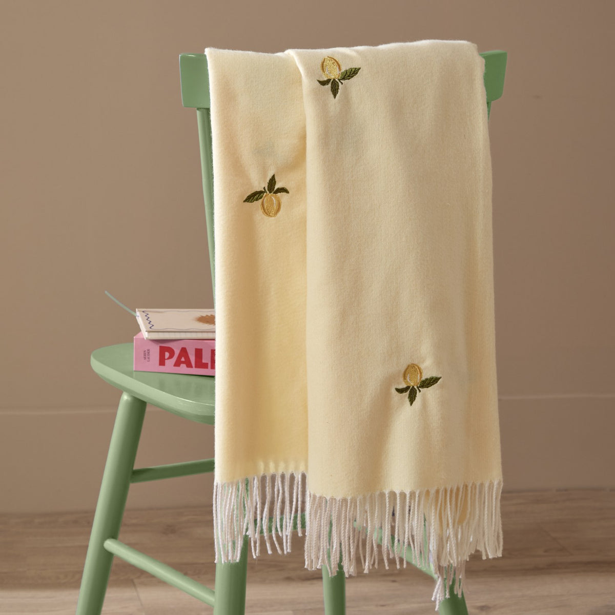 Fruits Knitted Polyester Throw in Zesty Lemon draped over a green chair, showing pale yellow fabric with embroidered lemon motifs and white tasselled fringe.