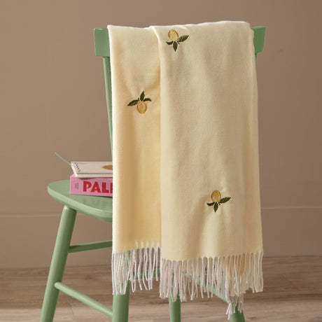 Fruits Knitted Polyester Throw in Zesty Lemon draped over a green chair, showing pale yellow fabric with embroidered lemon motifs and white tasselled fringe.