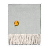 Sage green knitted polyester throw with an embroidered orange citrus motif and leafy sprig, finished with long cream tassels along one edge, shown folded on a plain white background.