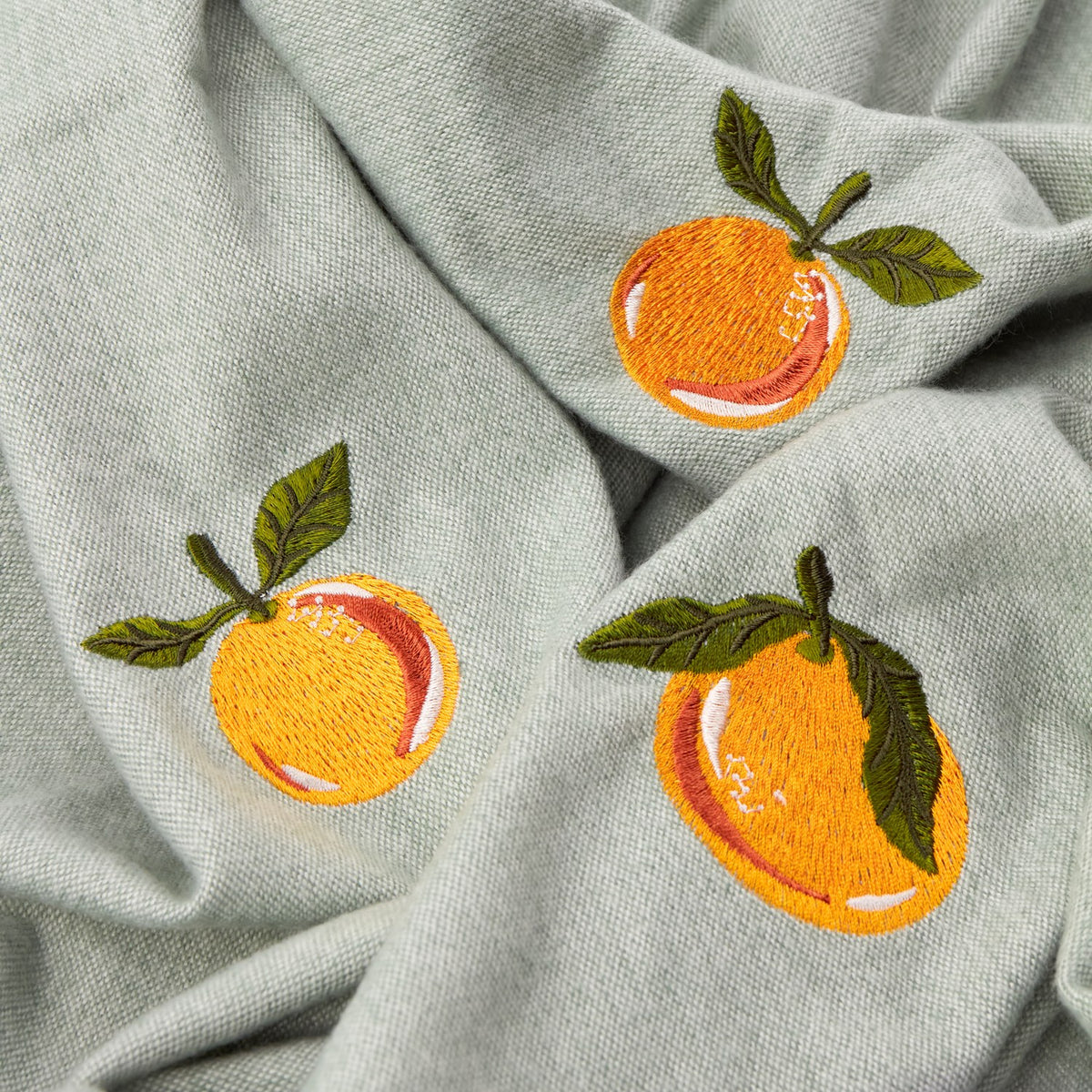 Close-up of the Fruits Knitted Polyester Throw in sage green, showing embroidered orange citrus motifs with green leafy sprigs on the knitted fabric.