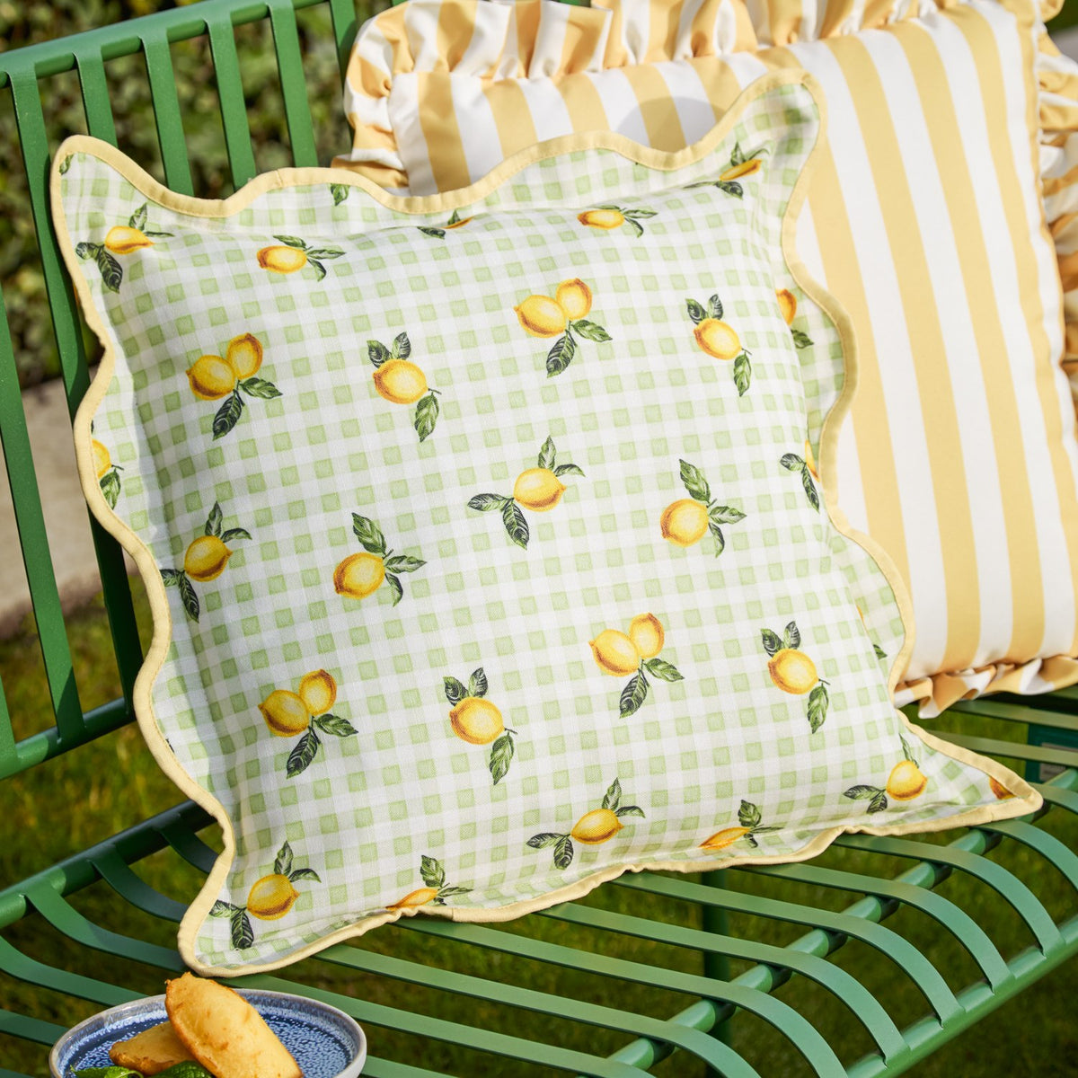 Fruits Gingham Polyester Cushion in Sage, a square scatter cushion with lemon print on pale green check fabric, scalloped edge and yellow piped trim, shown on a green garden bench.