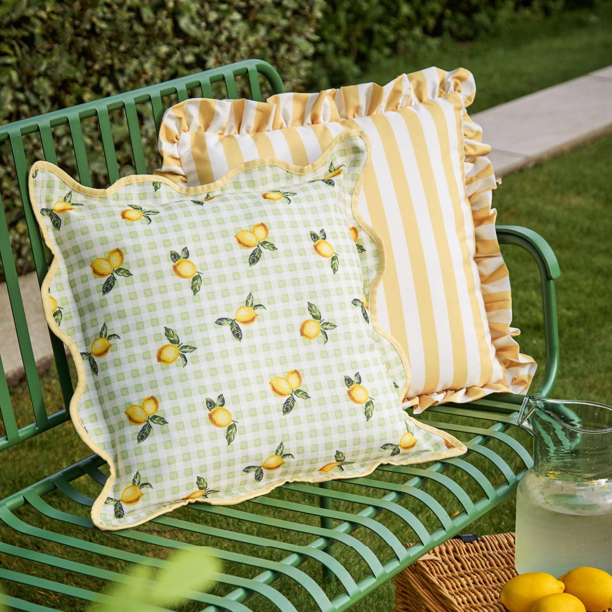 Fruits Gingham Polyester Cushion in Sage, a square scatter cushion with lemon print on pale green check, scalloped edge and yellow piped trim, shown on a green garden bench.