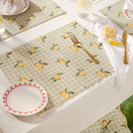 Fruits Gingham Polyester Placemat in sage on a dining table, woven fabric printed with lemons and leaves over a green and cream check, styled with cutlery, napkin, and a plate.