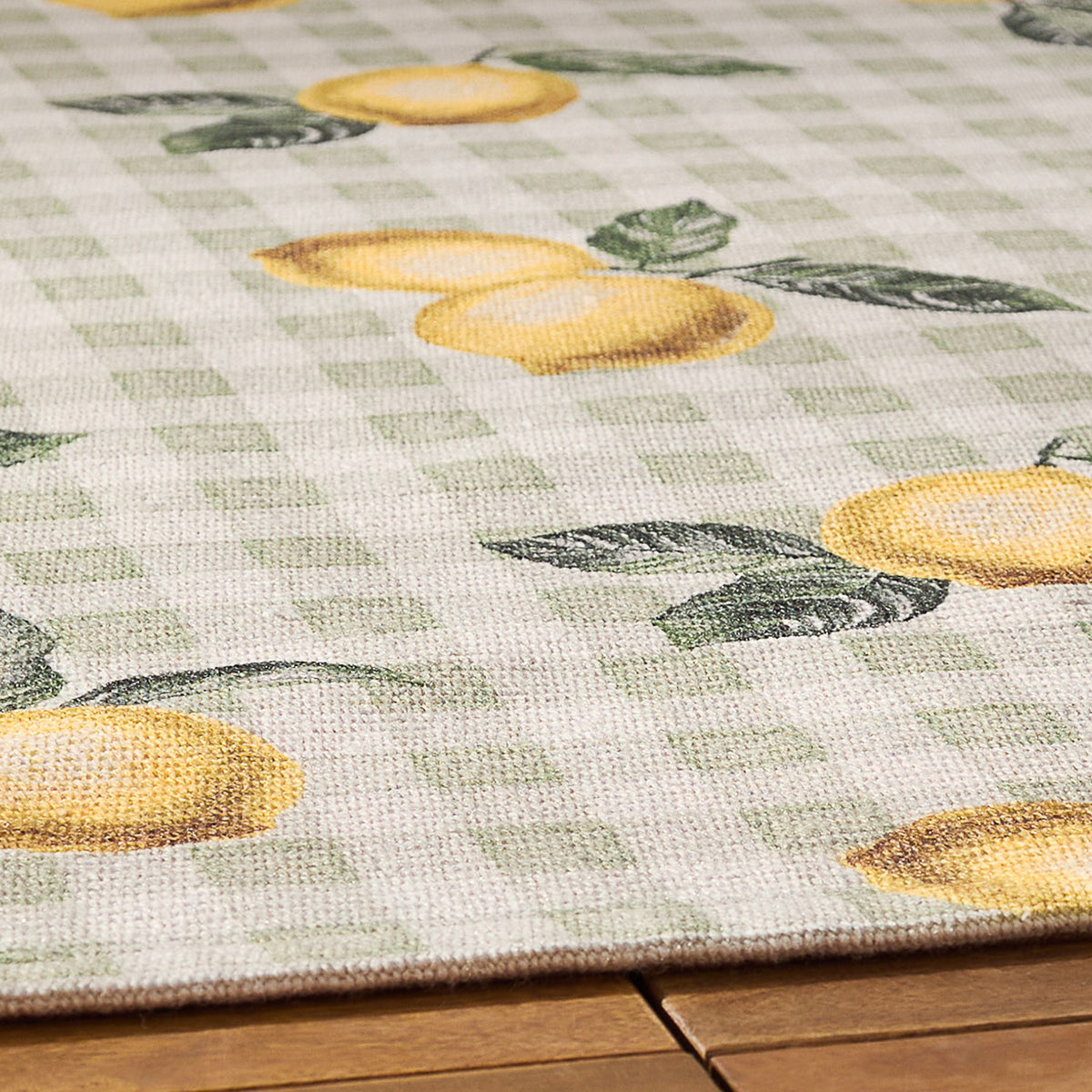 Close-up of the Fruits Gingham Polyester Rug in sage, showing a lemon and green leaf motif on a sage and cream check pattern, edge visible on a wooden floor.