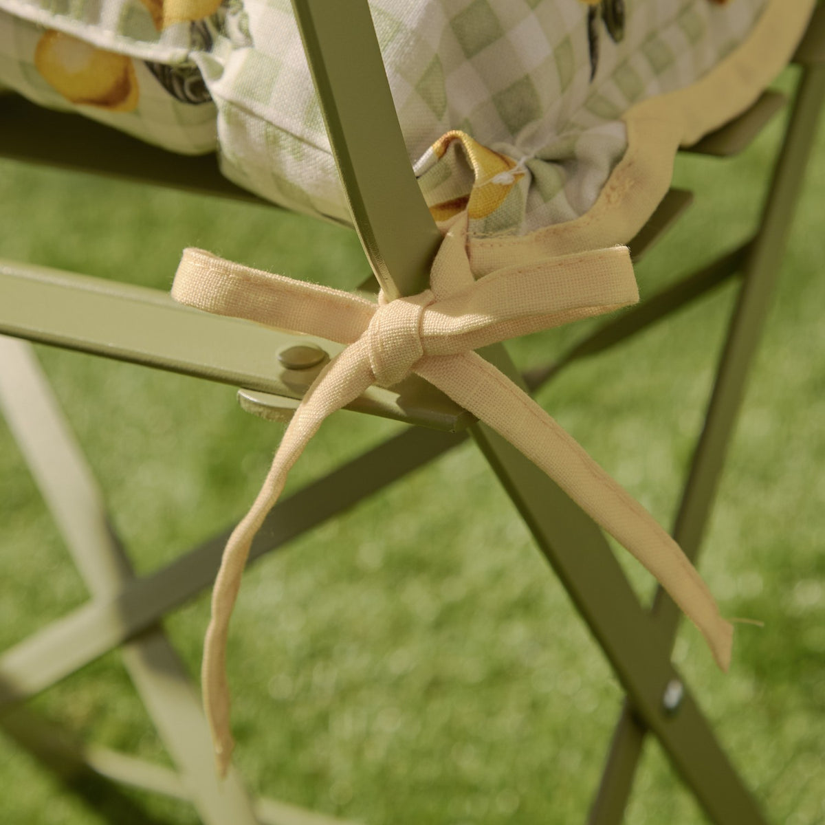 Close-up of the Fruits Gingham Polyester Seat Pad in sage, showing scalloped yellow piping and fabric ties knotted to a sage green garden chair frame.