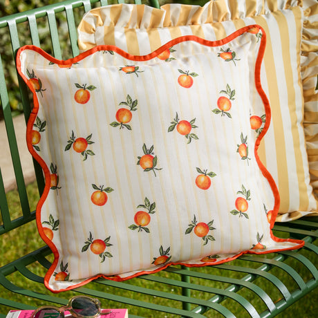 Square Fruits Striped Polyester Cushion in yellow, with scattered orange print on cream striped fabric and a scalloped orange piped edge, shown on a green garden chair.