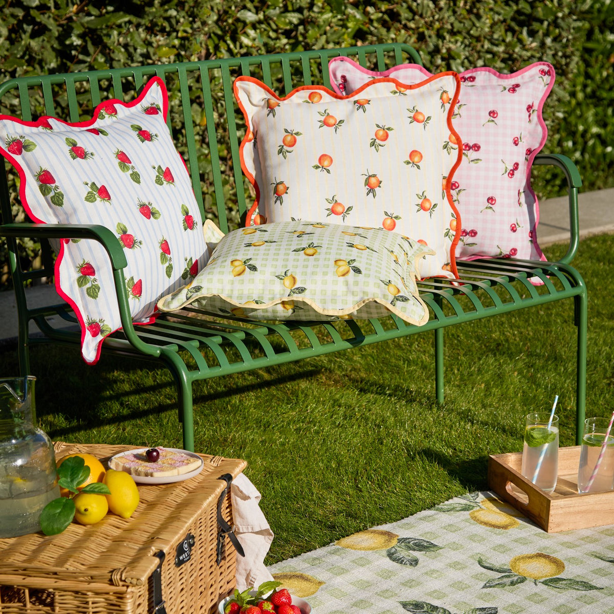 Square outdoor cushion with hand-painted orange print on cream and yellow striped fabric, finished with a scalloped edge and orange piped trim, styled on a garden bench.