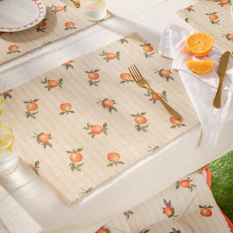 Fruits Striped Polyester Placemats in yellow with hand-painted orange motifs, set on an outdoor dining table with cutlery, napkin, and fresh orange slices.