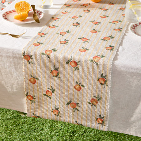Fruits Striped Polyester Table Runner in yellow, woven with cream and yellow stripes and scattered orange citrus print, shown laid over a white tablecloth with fringed ends.