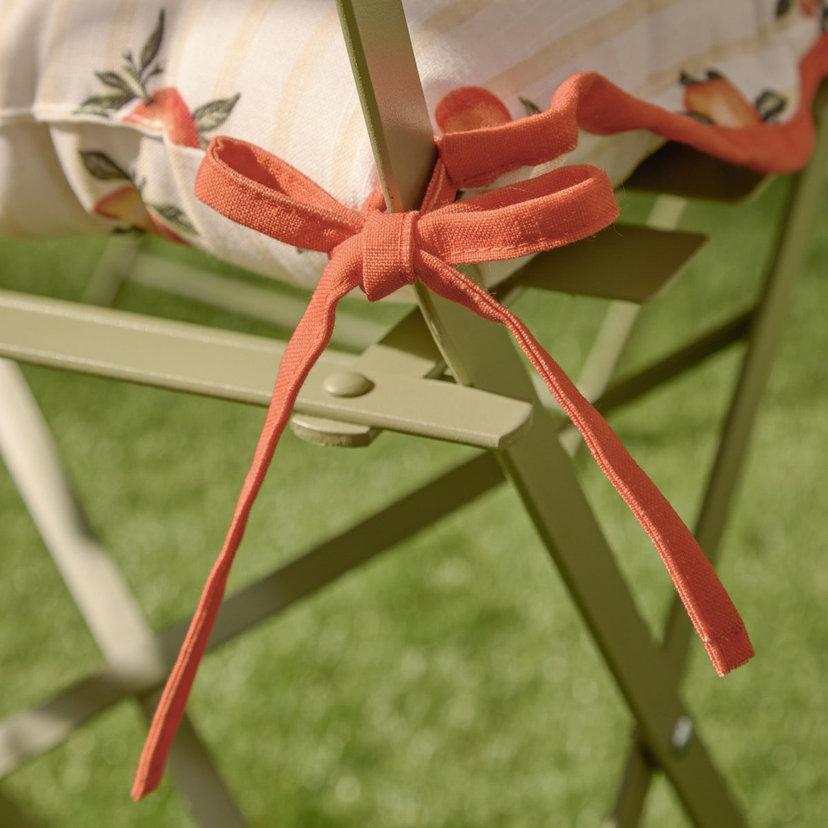Close-up of the Fruits Striped Polyester Seat Pad in yellow, showing rust-orange fabric ties knotted around a sage-green metal chair frame with scalloped trim visible.