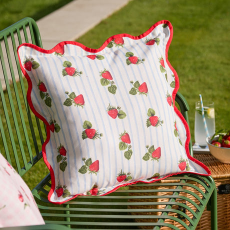 Square scatter cushion in blue striped polyester with a printed strawberry design and scalloped red piped edging, shown on a green garden chair outdoors.
