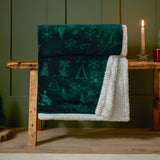 Festive Time Sherpa Woodland Polyester Throw in emerald green, draped over a wooden bench, showing embossed tree and snowflake pattern with white sherpa reverse.