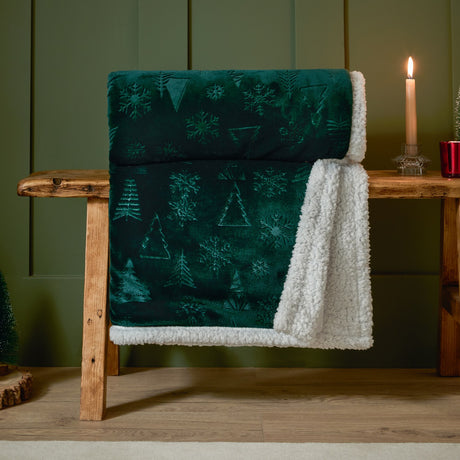 Festive Time Sherpa Woodland Polyester Throw in emerald green, draped over a wooden bench, showing embossed tree and snowflake pattern with white sherpa reverse.