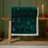 Festive Time Sherpa Woodland Polyester Throw in emerald green, folded over a wooden bench with embossed tree and snowflake pattern and white sherpa reverse visible, styled near a lit candle.