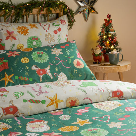 Emerald reversible duvet cover set on a bed, printed with gingerbread houses, Santa faces, candy canes, wreaths and star biscuits, styled in a bedroom with a lit Christmas tree.