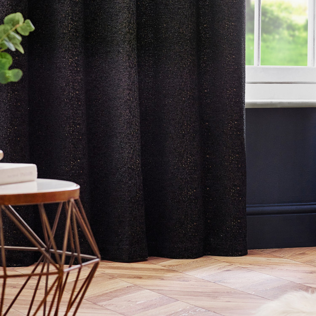 Galaxy Chenille fully lined eyelet curtains in black with subtle gold flecks, shown in soft folds beside a window in a living room with wooden flooring.