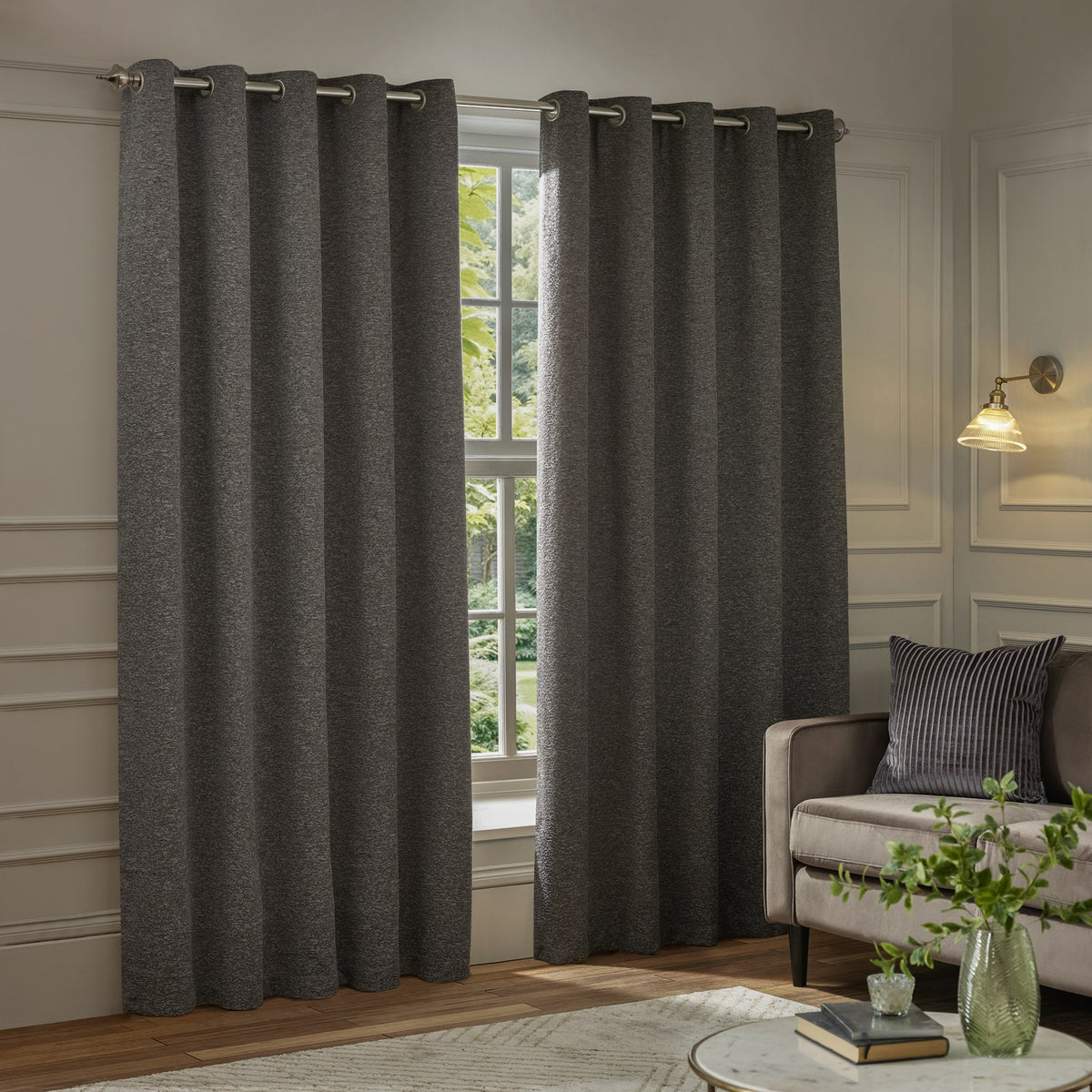Galaxy Chenille Fully Lined Eyelet Curtains in charcoal, hung on a metal pole beside a window in a living room with a sofa and side table.