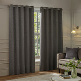 Galaxy Chenille Fully Lined Eyelet Curtains in charcoal, hung on a metal pole beside a window in a living room with a sofa and side table.