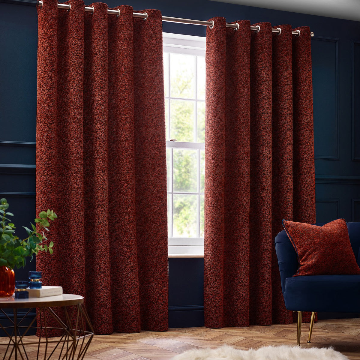 Galaxy Chenille Fully Lined Eyelet Curtains in copper, textured rust-toned fabric with silver eyelet heading, hung on a pole in a navy-walled living room beside a window.