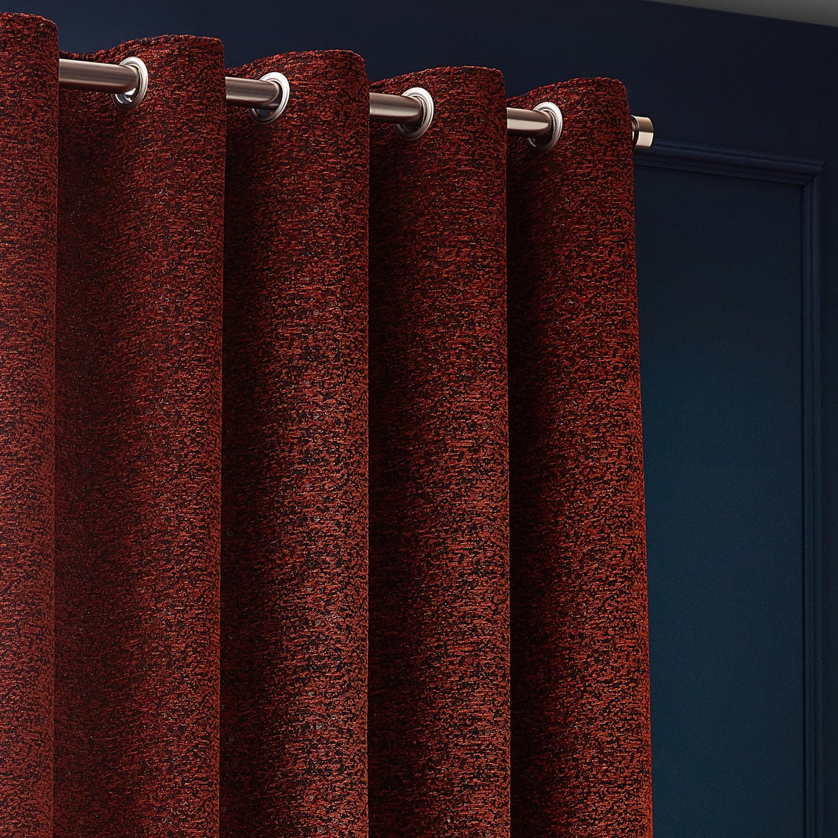 Galaxy Chenille Fully Lined Eyelet Curtains in copper, close-up of textured rust fabric with brushed metal eyelets on a pole against a dark blue wall.