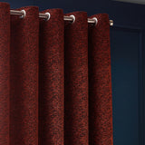 Galaxy Chenille Fully Lined Eyelet Curtains in copper, close-up of textured rust fabric with brushed metal eyelets on a pole against a dark blue wall.