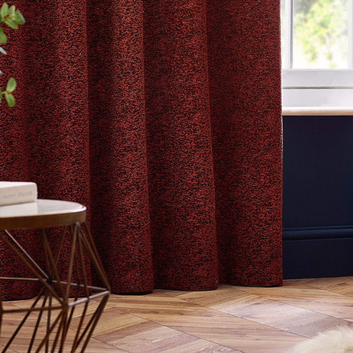 Galaxy Chenille Fully Lined Eyelet Curtains in copper, hung at a window in a living room with dark blue panelling and wood flooring, showing textured rust-toned fabric with golden flecks.