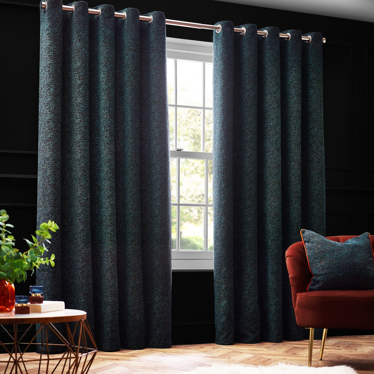 Galaxy Chenille Fully Lined Eyelet Curtains in emerald green with subtle gold flecks, shown hanging on a rod in a dark living room beside a window and rust-coloured armchair.