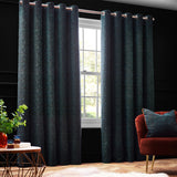 Galaxy Chenille Fully Lined Eyelet Curtains in emerald green with subtle gold flecks, shown hanging on a rod in a dark living room beside a window and rust-coloured armchair.